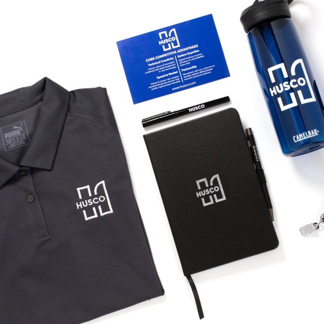 An overhead shot of a polo with the husco logo on it next to a husco branded notebook, water bottle, and pen.